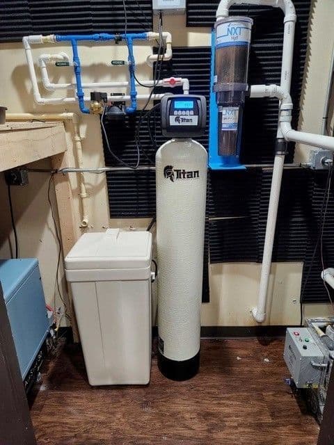 Titan whole-home water treatment system with tanks, pipes, and control panel against a wall.