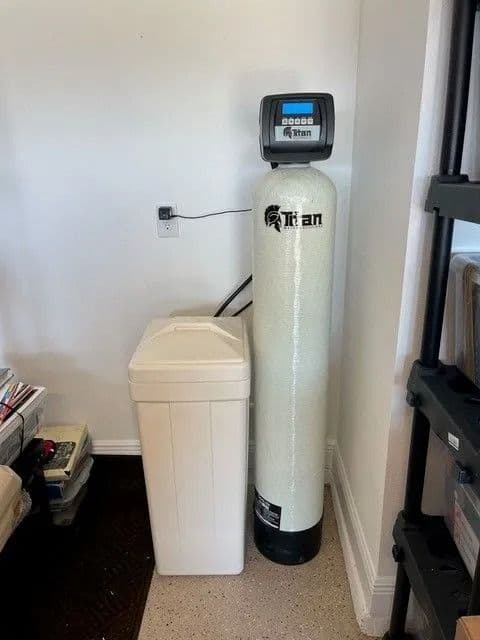 Water softener system in a corner. A tall white tank with a control panel stands next to a white salt storage tank.