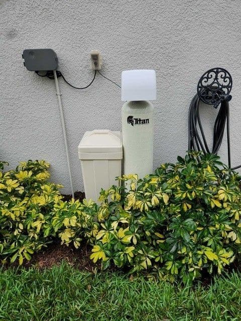 A Titan whole-home water treatment system against a stucco wall, surrounded by green and yellow shrubs.