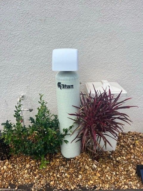 Titan water filter beside a small plant, on a gravel bed, against a light wall.