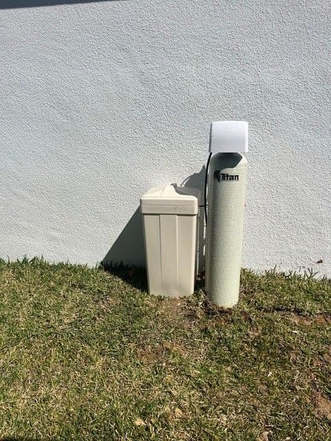 Titan whole-home water treatment system next to a stucco wall, with a small salt tank on the left and a tall filtration tank on the right, outdoors.