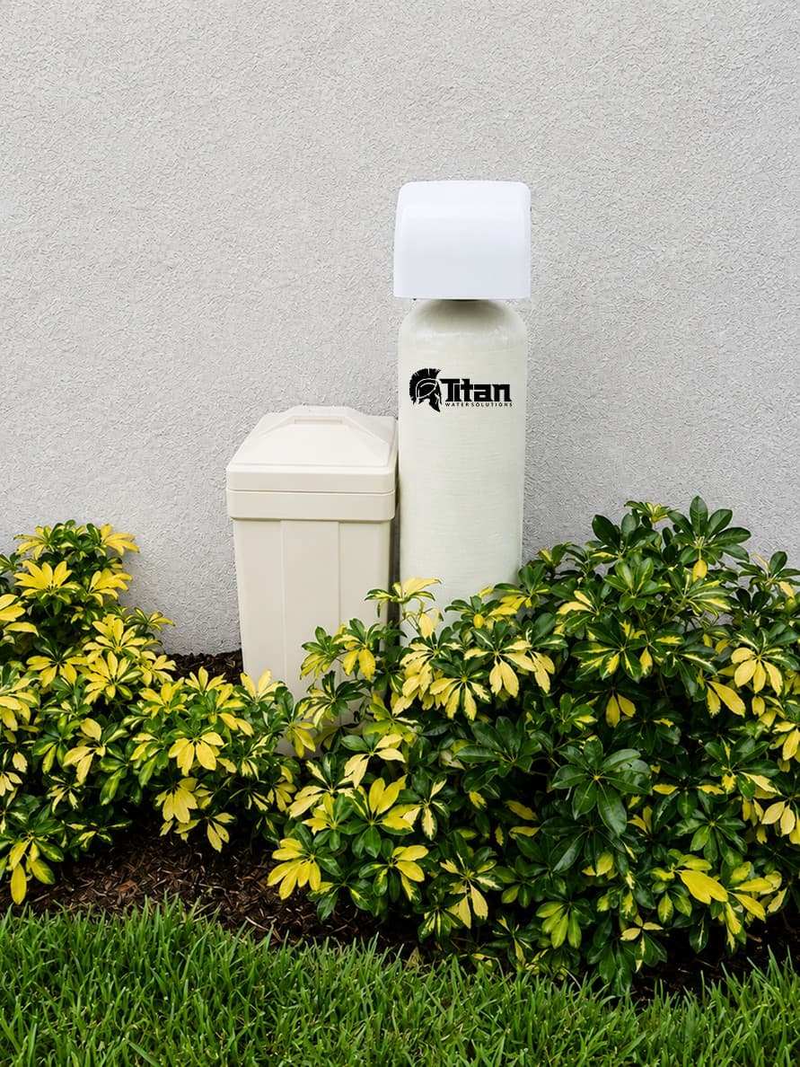 A Titan water softener and salt tank installed outdoors, surrounded by shrubs