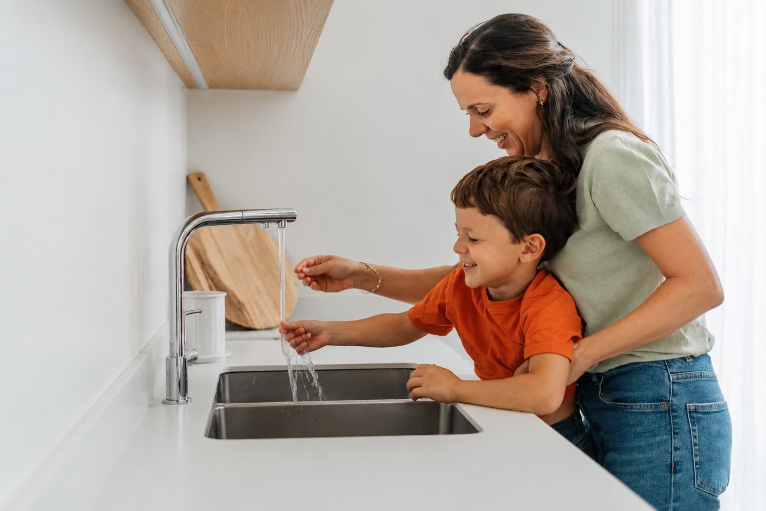 A mother and young son washing produce together at a bright kitchen sink